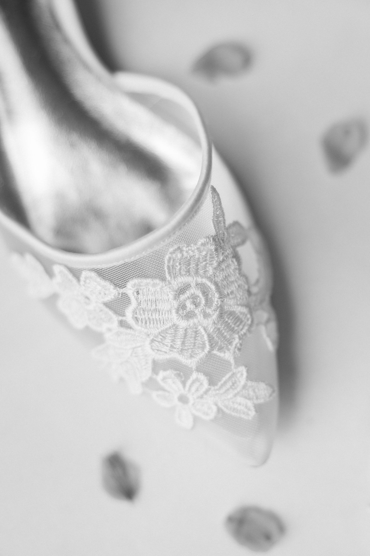A delicate white ballet slipper with lace floral embroidery detail shown in soft focus black and white photography.