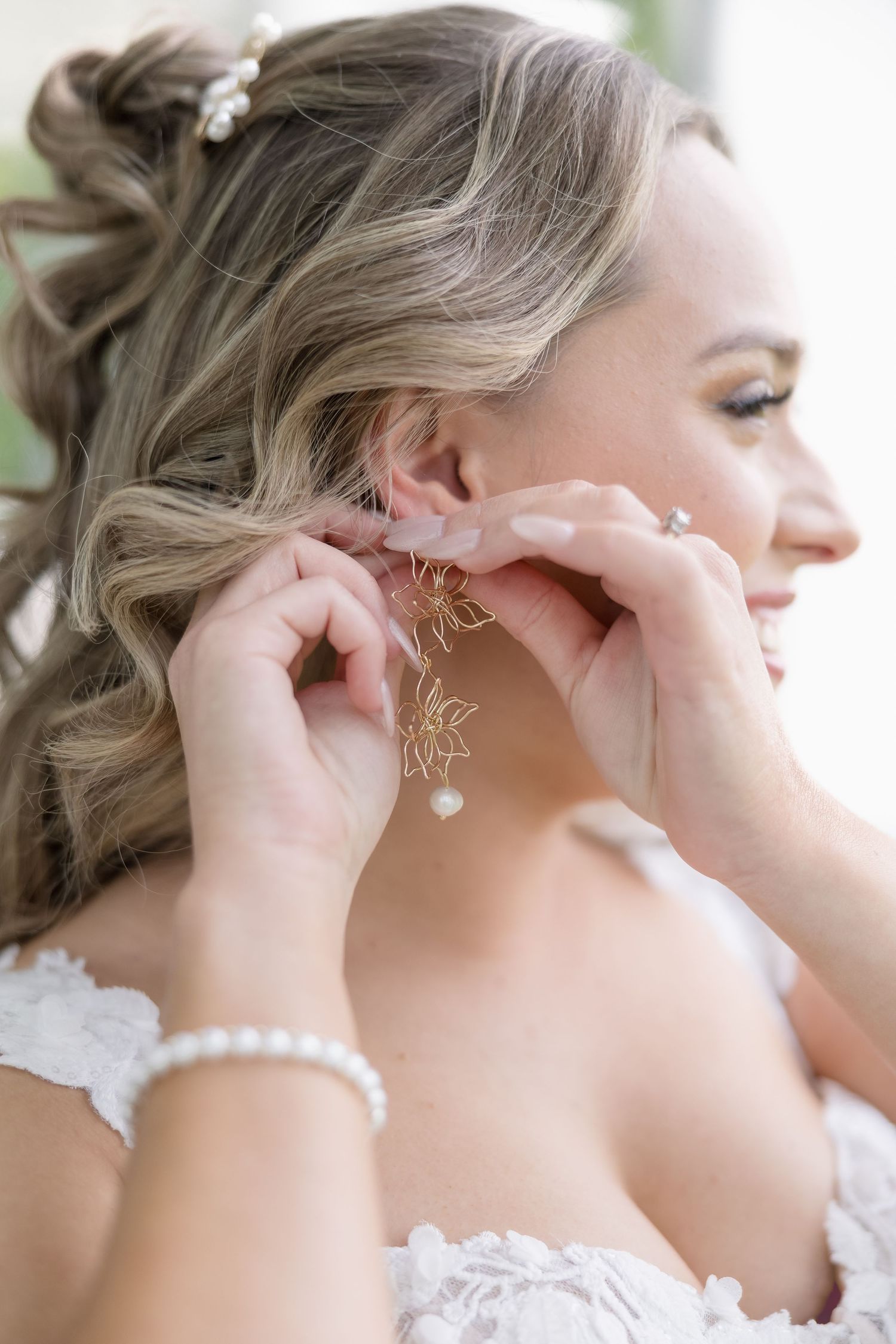 Hands putting on delicate pearl and crystal earrings with an elegant white lace dress and pearl bracelet in view.