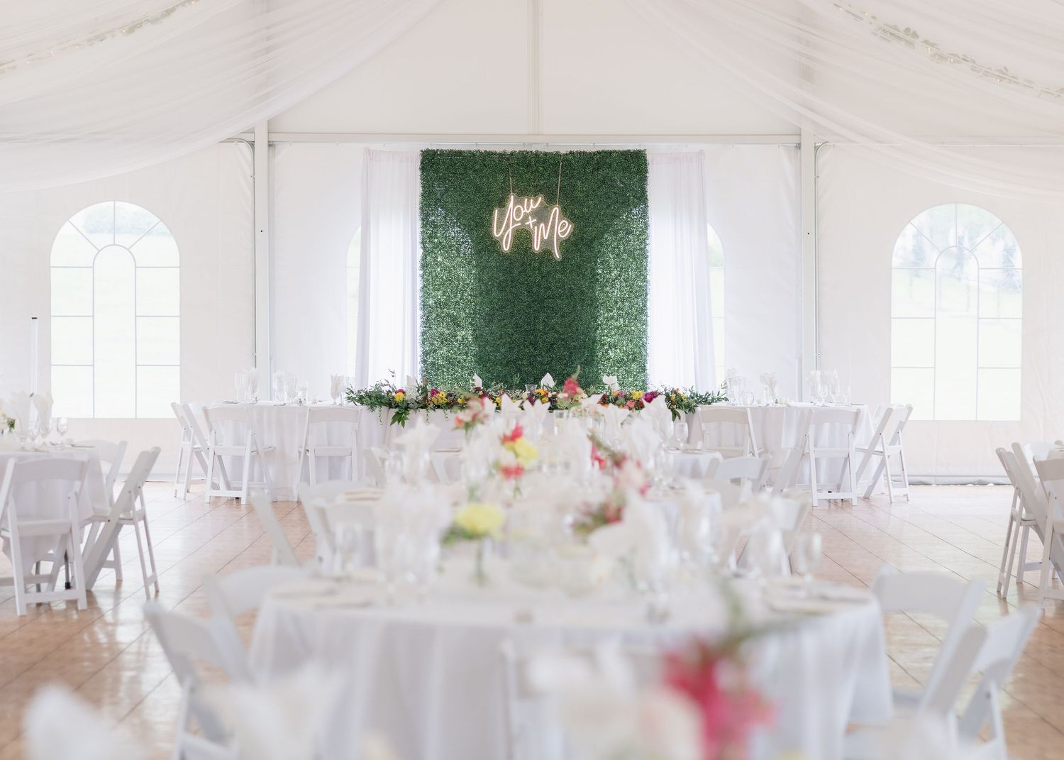 White wedding reception tent with round tables and an elegant green floral backdrop at the front of the venue.