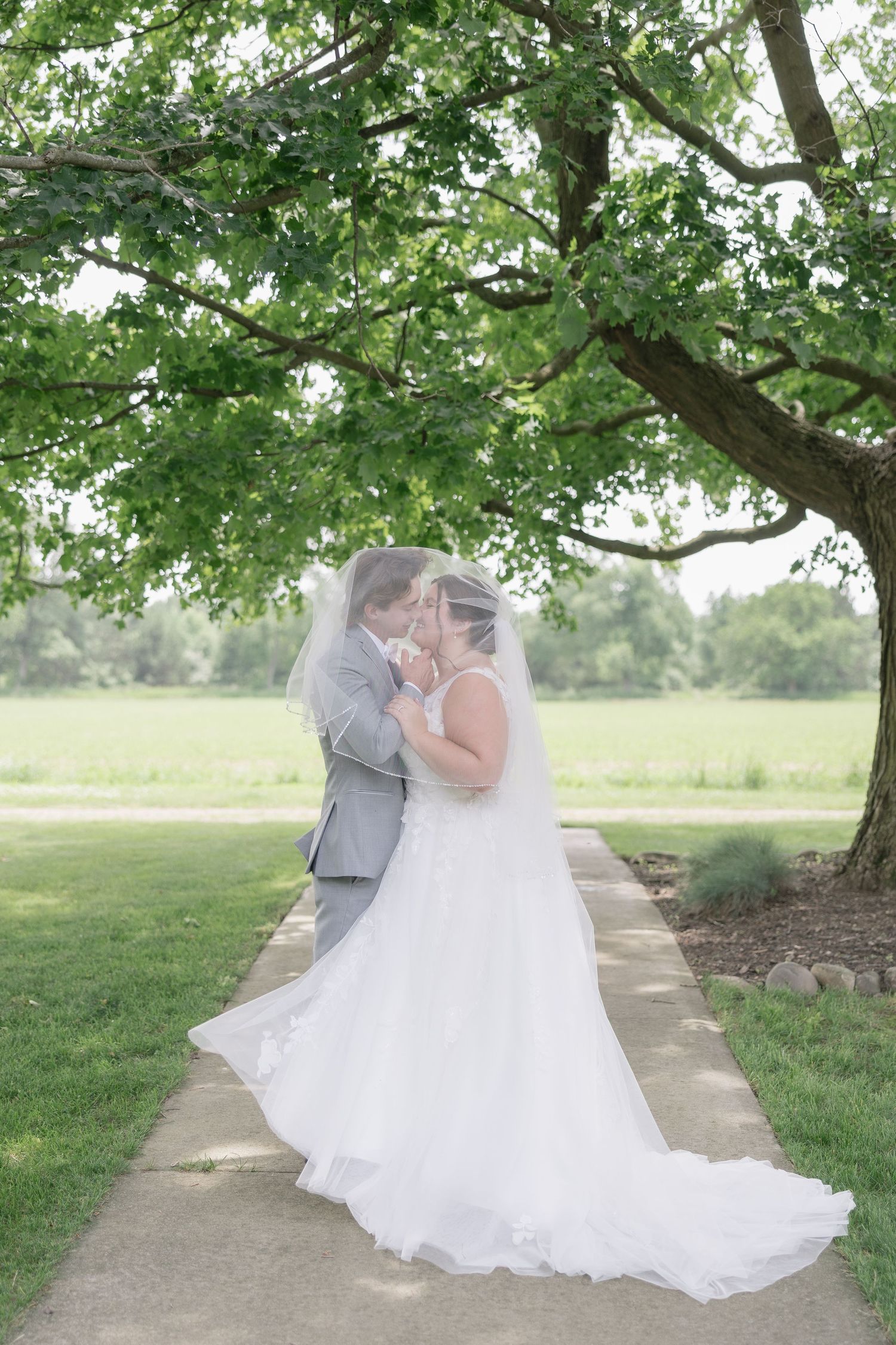 A couple in wedding attire sharing an intimate moment under a lush green tree along a garden path.