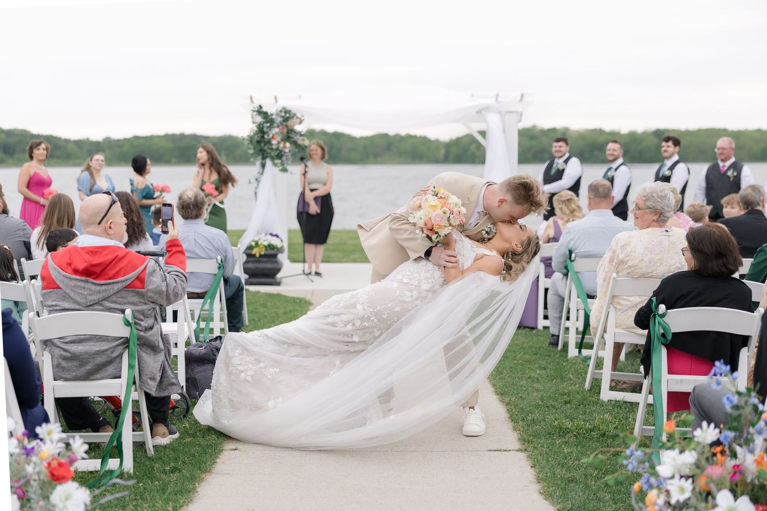 A romantic moment during an outdoor waterfront wedding ceremony with guests seated on white chairs and colorful floral arrangements.