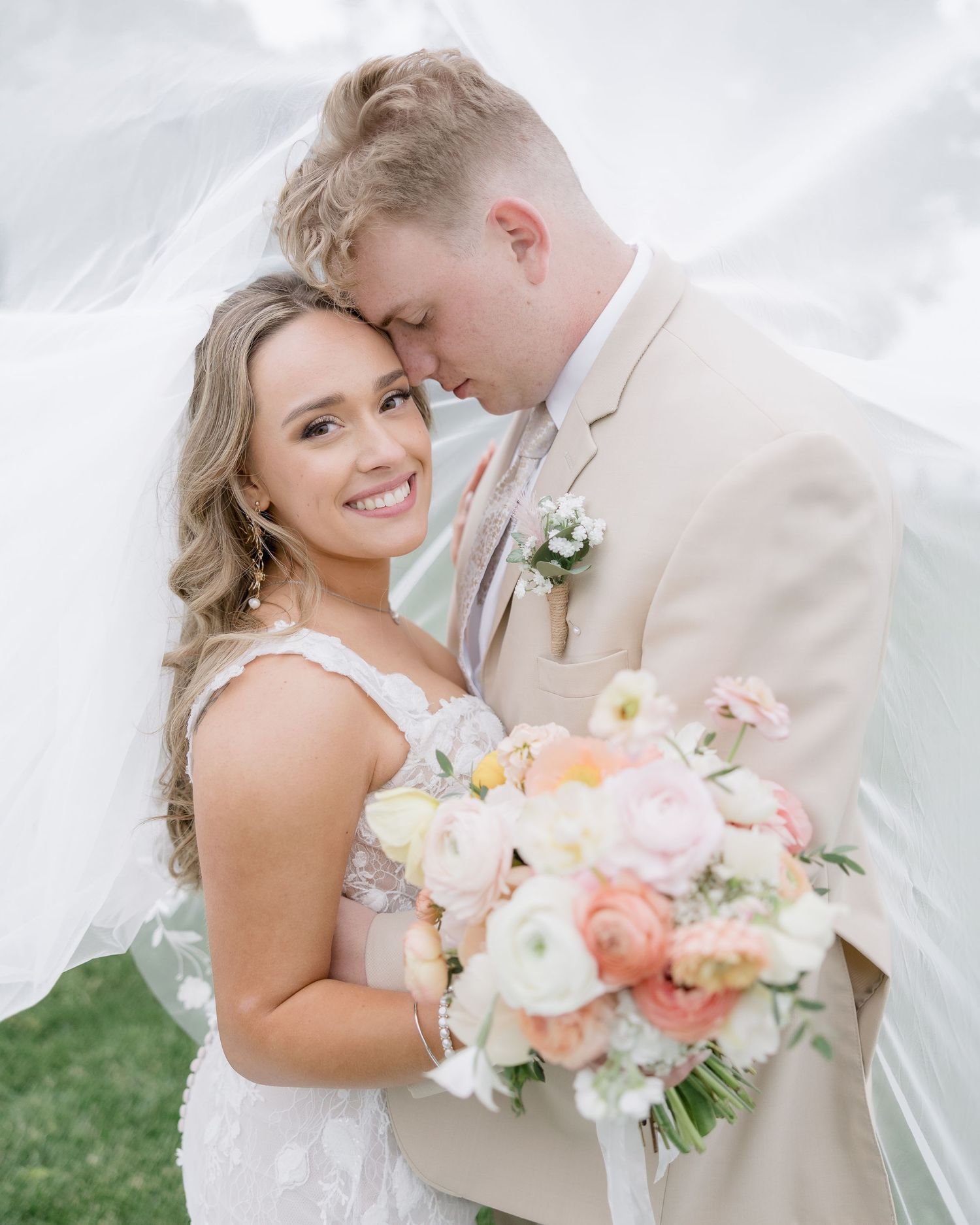 Romantic couple in wedding attire share intimate moment with bride holding lush pink and peach floral bouquet.