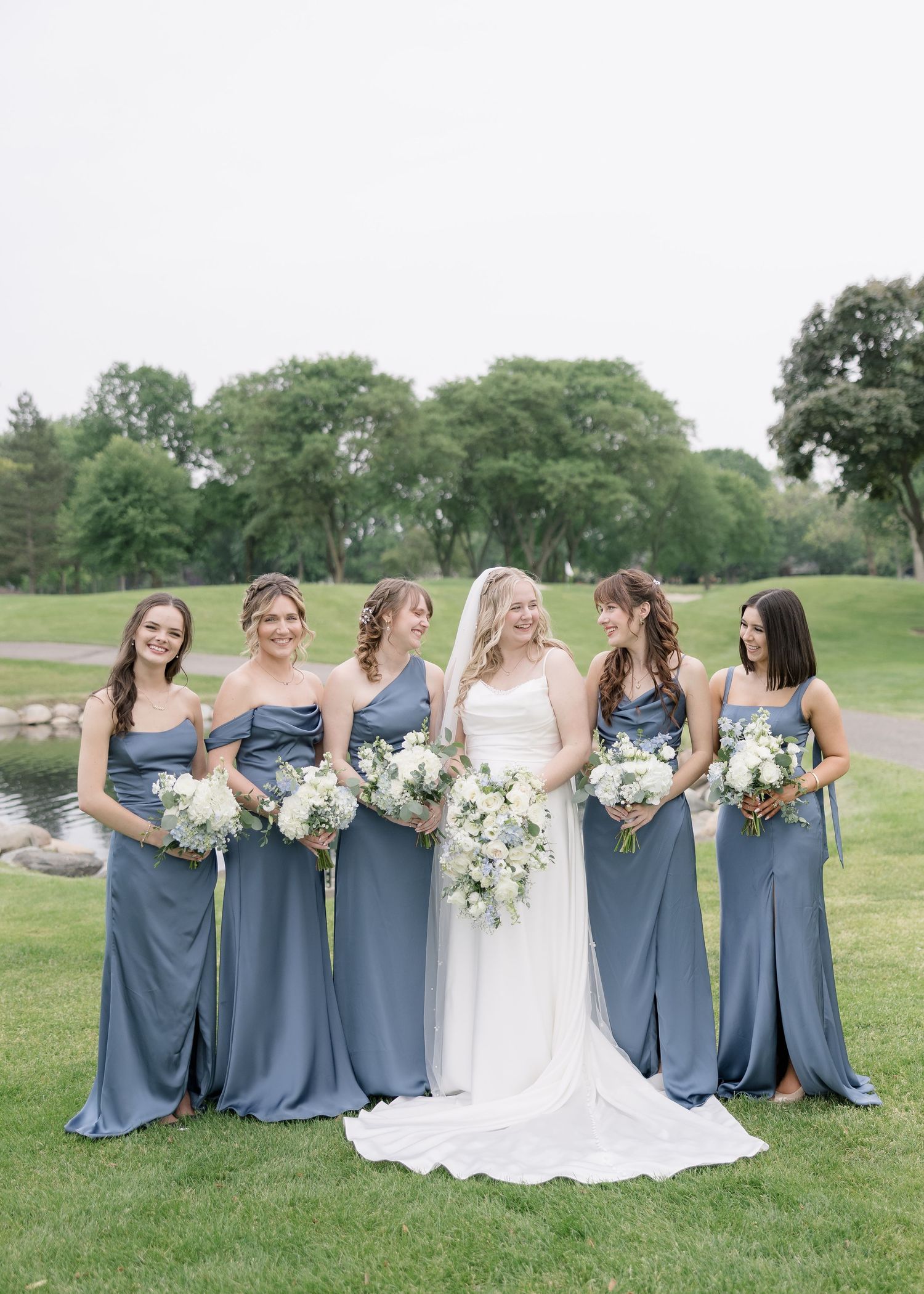 Wedding party wearing dusty blue bridesmaid dresses pose together on grassy hill at outdoor venue.