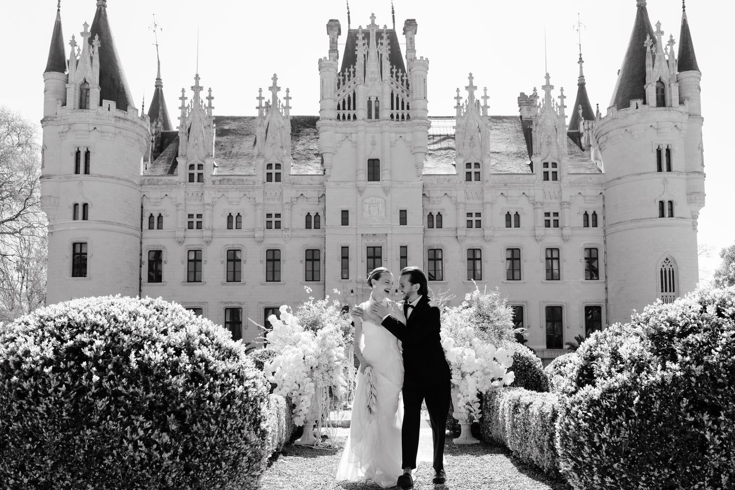A romantic wedding couple embraces in front of a grand castle with gothic spires and beautiful flowering bushes.