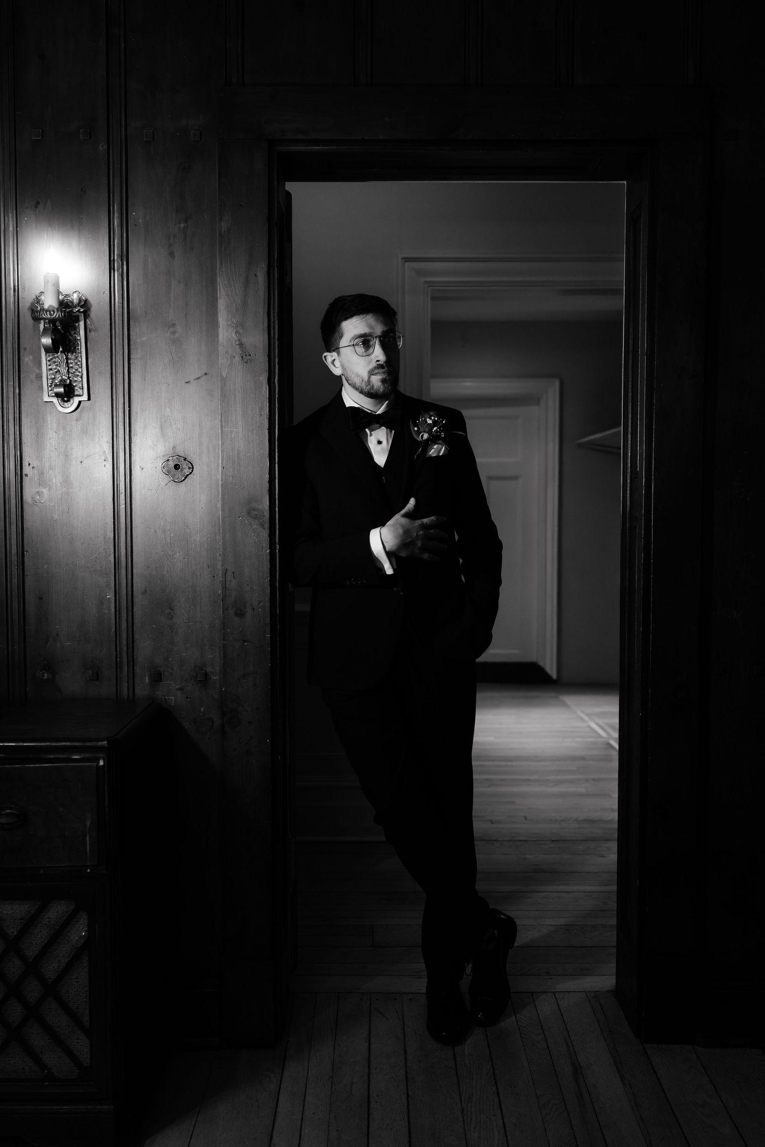 A dapper groom in a black tuxedo and bow tie leans against a doorframe in a dramatic black and white portrait.