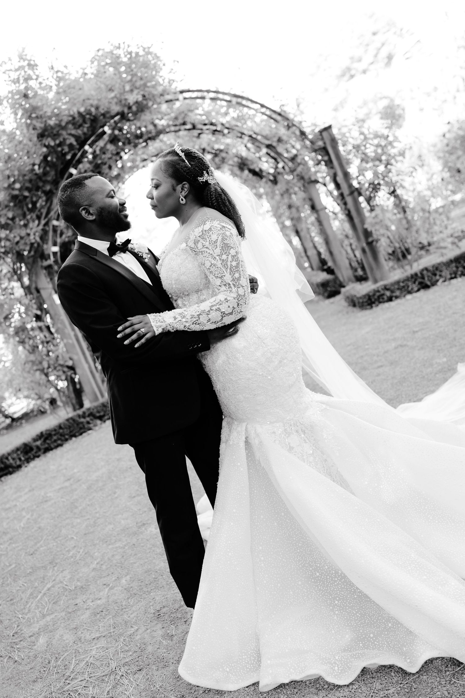 A bride and groom share a romantic moment under a garden archway in an elegant black and white wedding photo.