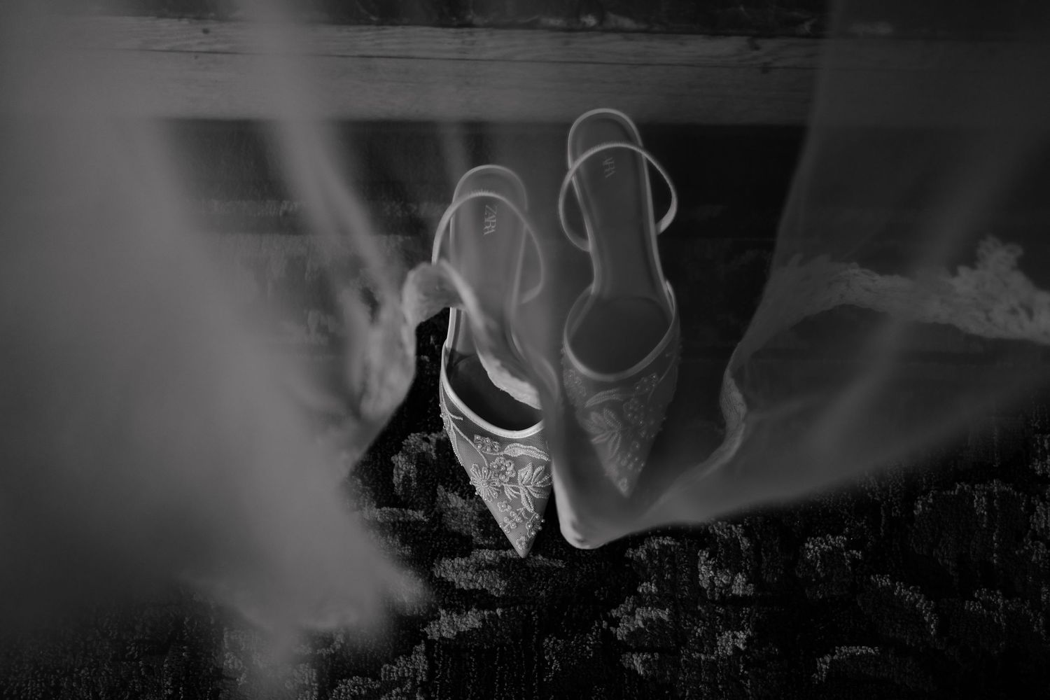 Black and white photo of elegant bridal heels with lace detail resting beneath a flowing wedding veil.
