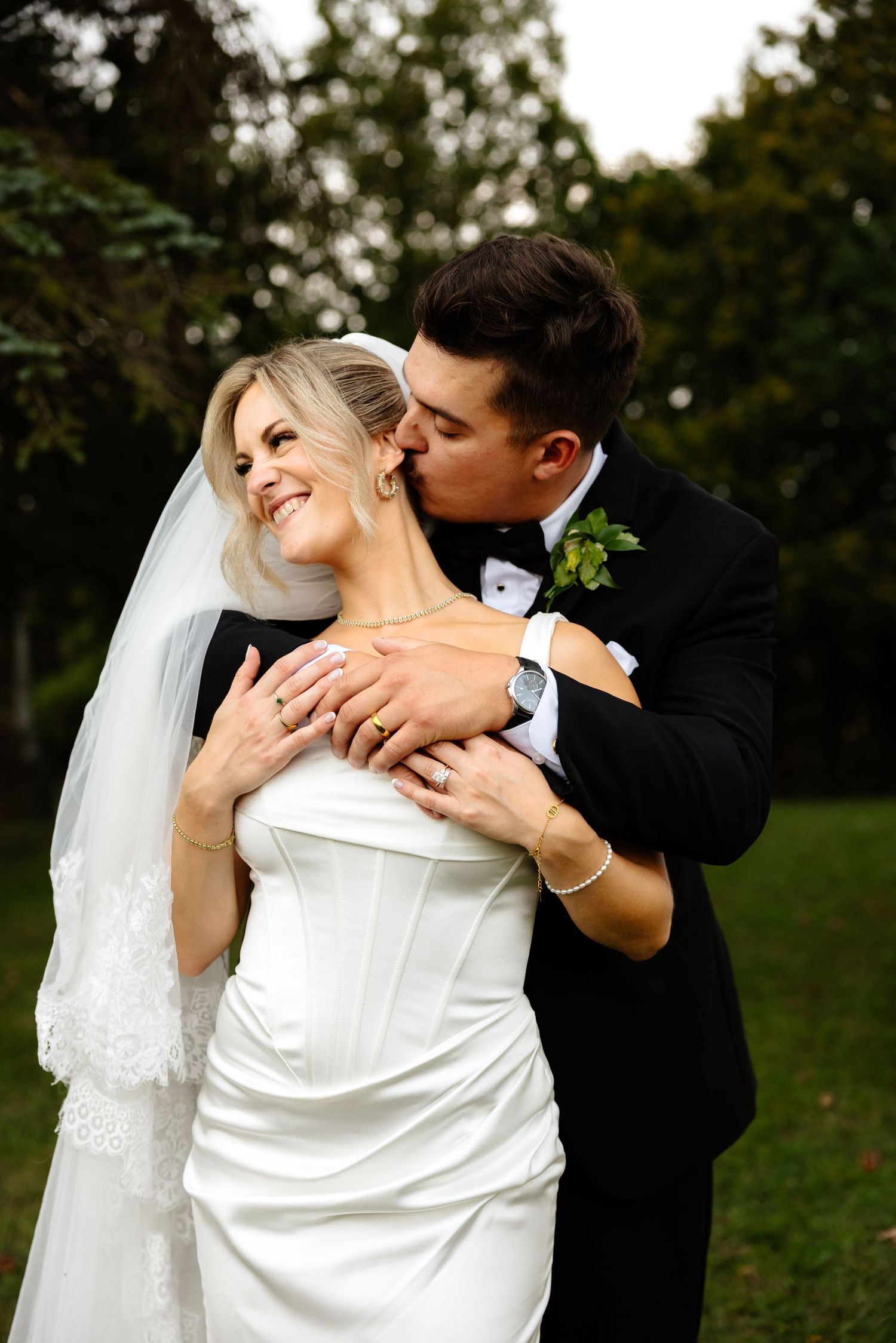 Groom in black tuxedo kisses bride on cheek as she smiles, wearing white strapless gown and veil outdoors.