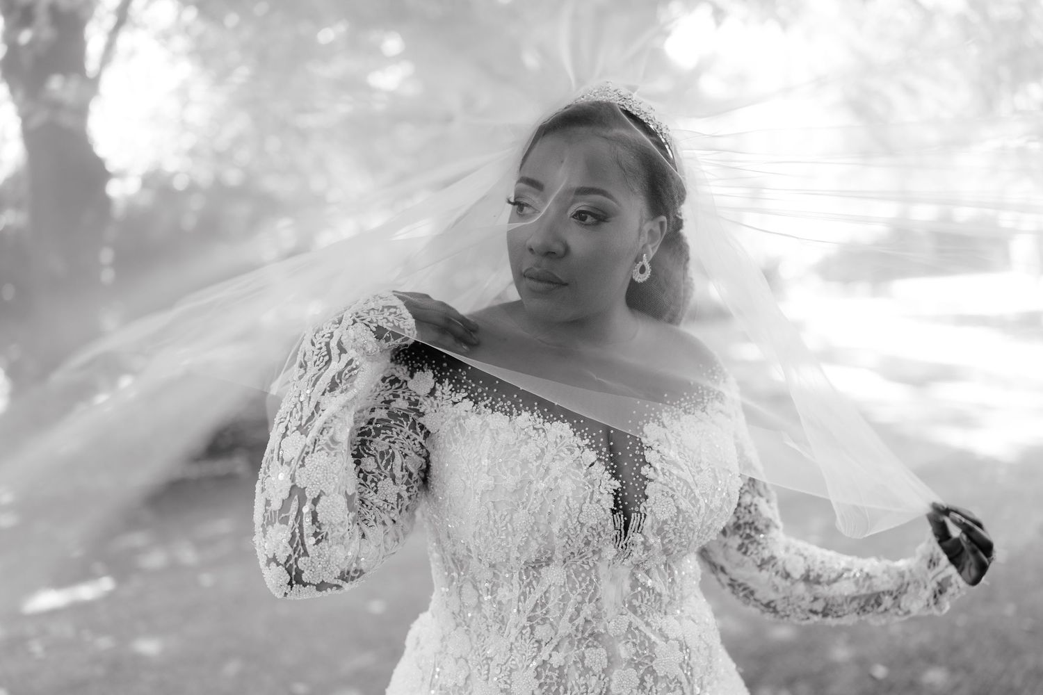 Black and white photo of a bride in lace gown with flowing veil, posing outdoors on her wedding day.