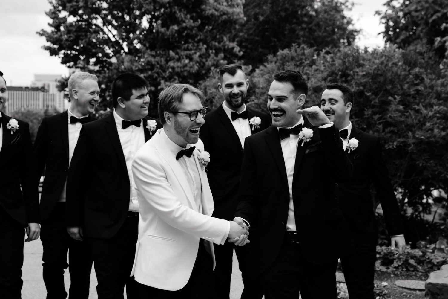 Groom in white tuxedo shaking hands with groomsmen in black tuxedos, laughing outdoors at a wedding.