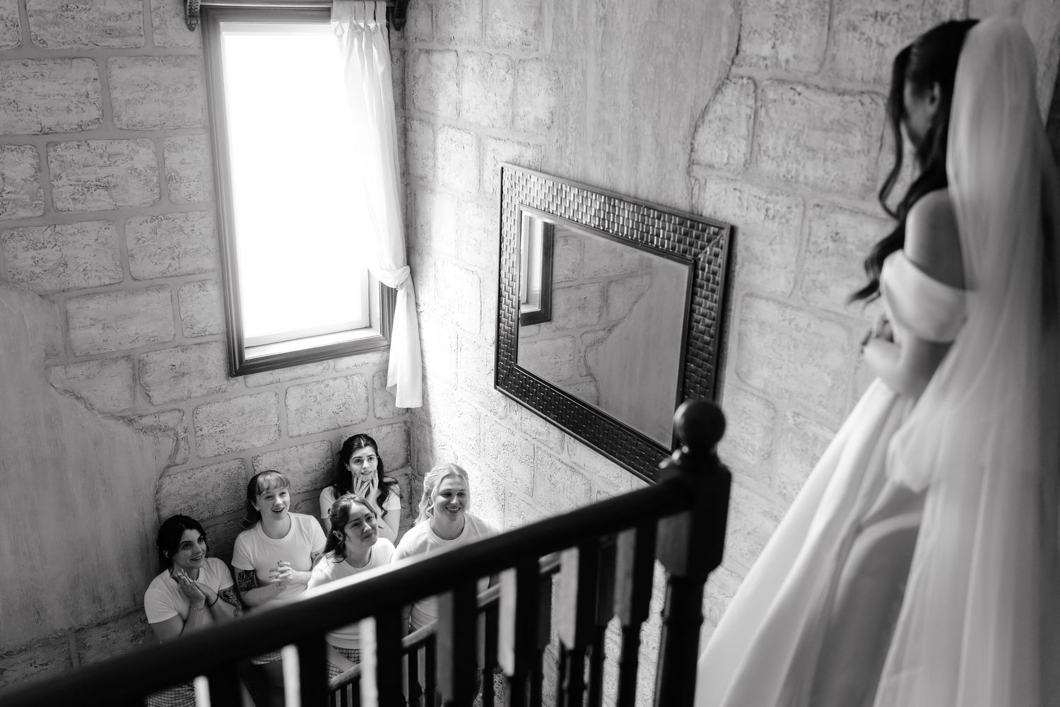 Bride in veil stands at staircase railing as bridesmaids look up admiringly in black and white wedding photo.