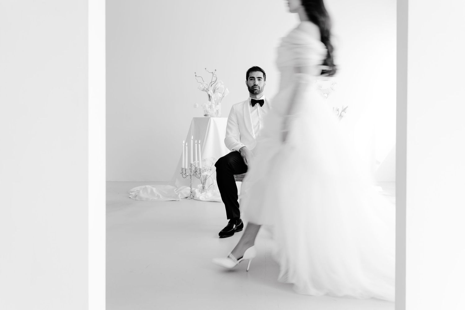 Black and white wedding photo of groom in tuxedo standing while bride in white gown walks toward him in bright studio.
