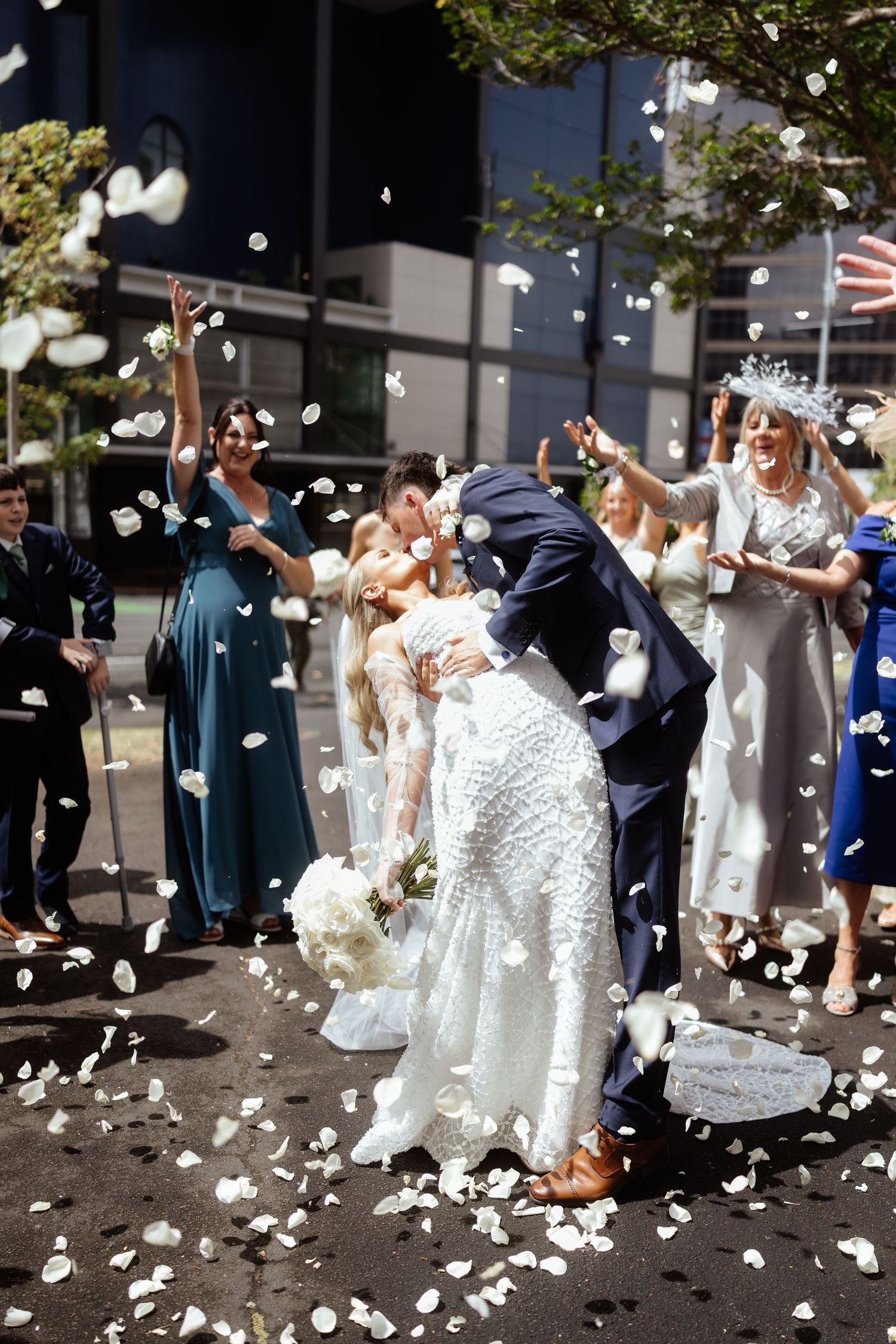 Wedding guests toss white flower petals at a couple during a joyful outdoor celebration in front of a building.