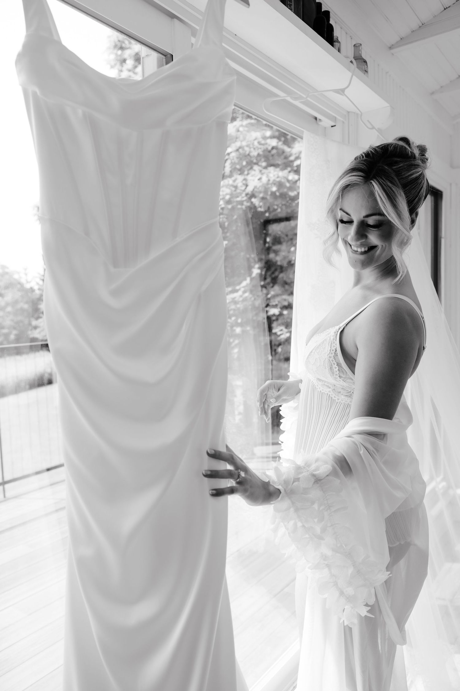 Black and white photo of a smiling bride in a white gown holding her wedding dress near a bright window.