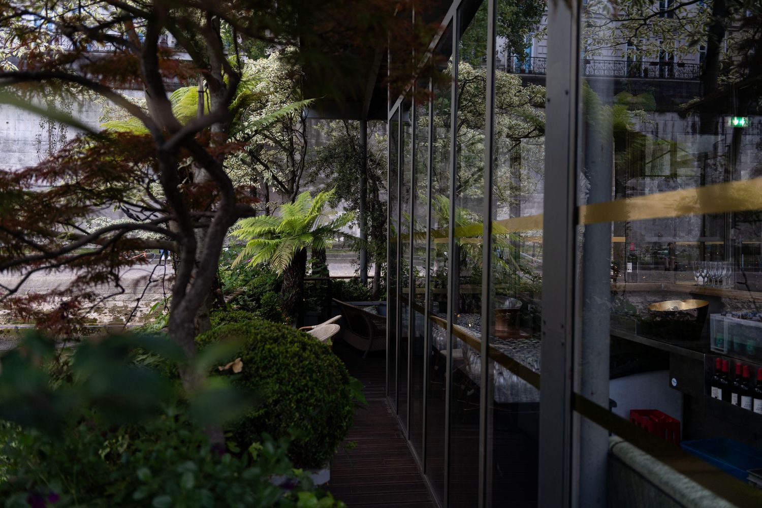 Glass-walled restaurant or cafe interior with lush garden landscaping and trees visible through reflective windows.