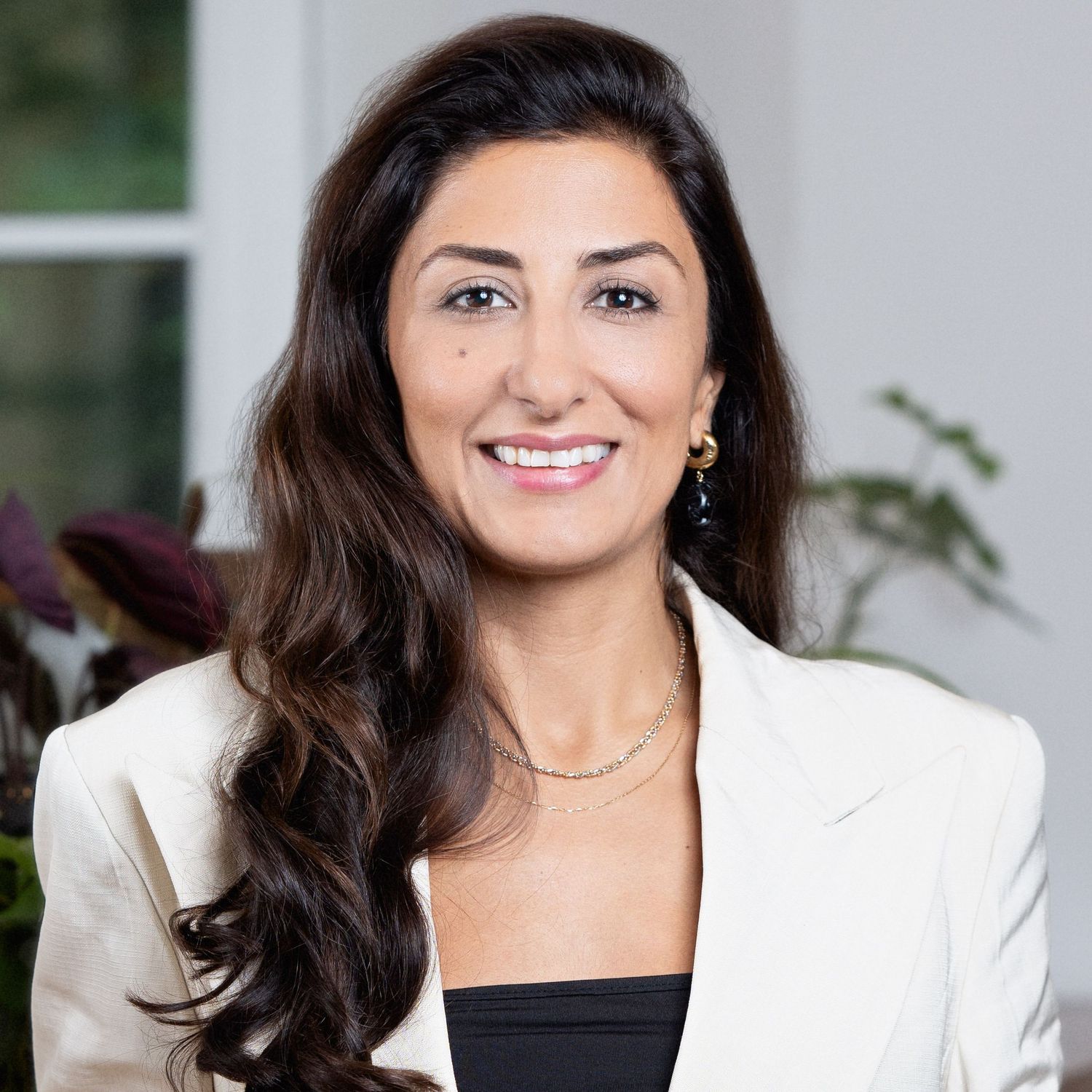 Professional headshot of a smiling person with long dark hair wearing a white blazer and black top against a blurred background.