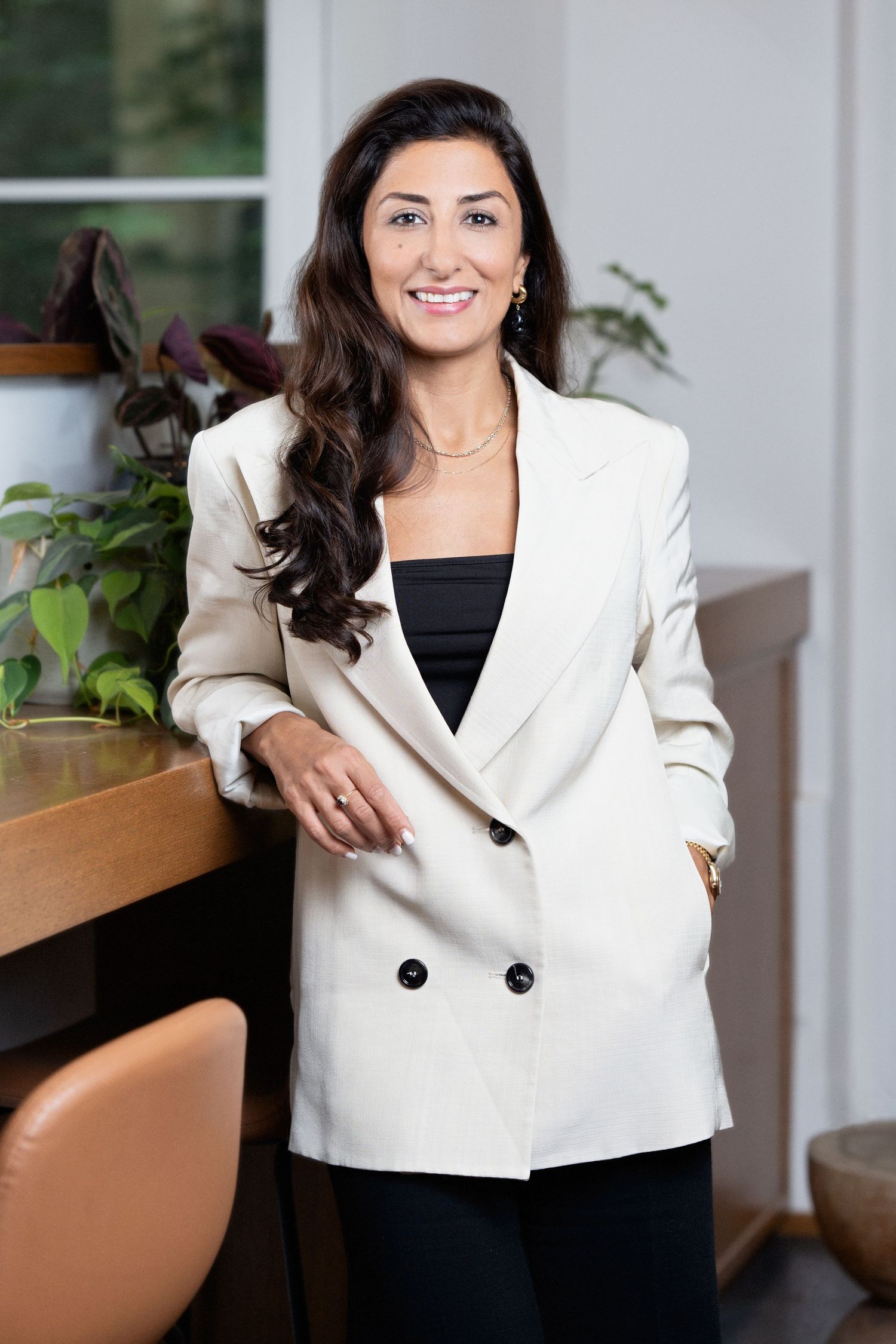 Professional portrait in modern office setting featuring business attire of white blazer and black top amid indoor plants.