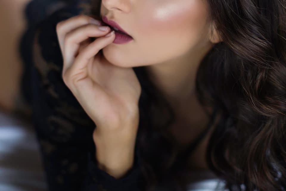 A woman with dark curly hair and burgundy lipstick holds her fingers to her lips in a thoughtful, elegant pose.