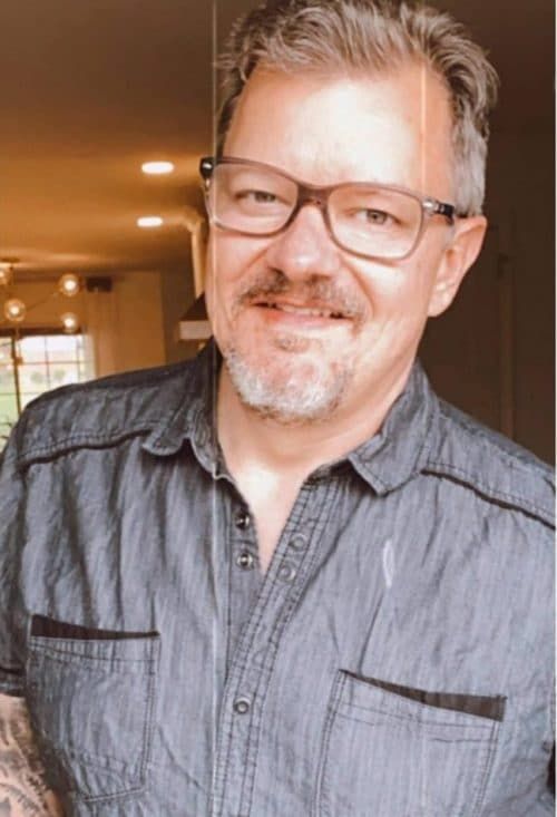 A person wearing glasses and a gray chambray button-down shirt smiles warmly at the camera in an indoor setting.