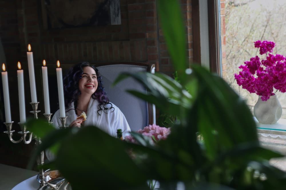 A cozy dining scene with candlelight, vibrant pink flowers, and greenery by a bright window.