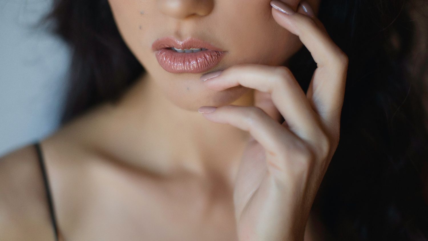 Close-up portrait of a young woman with glossy lips and dark hair touching her face gently.