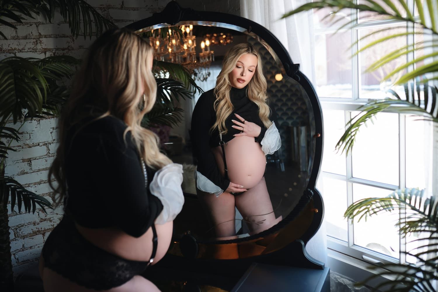 Pregnant woman in black lingerie admiring her baby bump in a large round mirror surrounded by lush green plants.