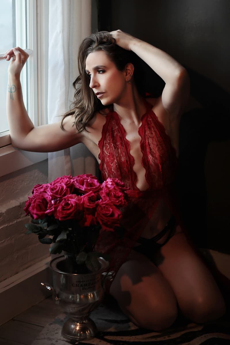 A sensual boudoir photoshoot featuring red lace lingerie and a bouquet of vibrant red roses by a window.