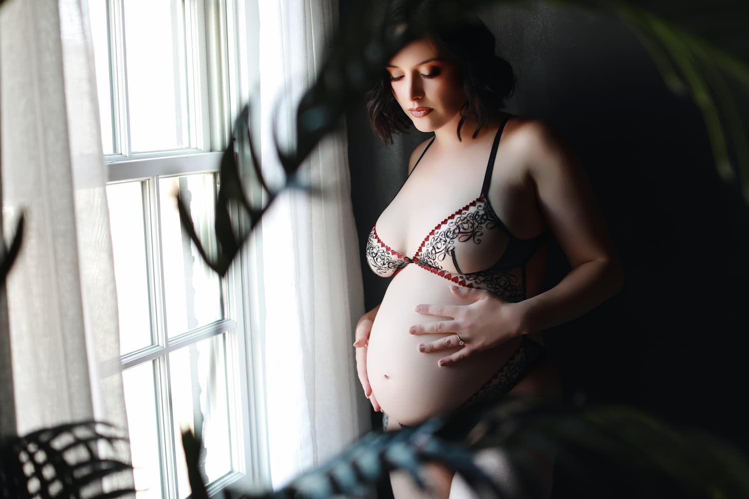 Pregnant woman in red lace lingerie cradling her baby bump by a bright window with soft natural light.