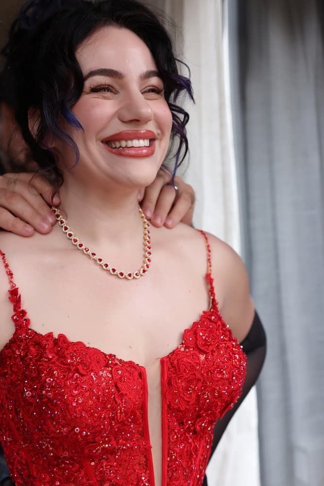 A person wearing a red lace formal dress and gold necklace smiles brightly near a window.