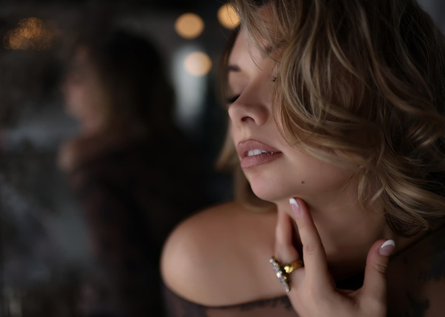A woman with blonde hair touches her chin, wearing a gold ring, in a moody, bokeh-lit portrait.
