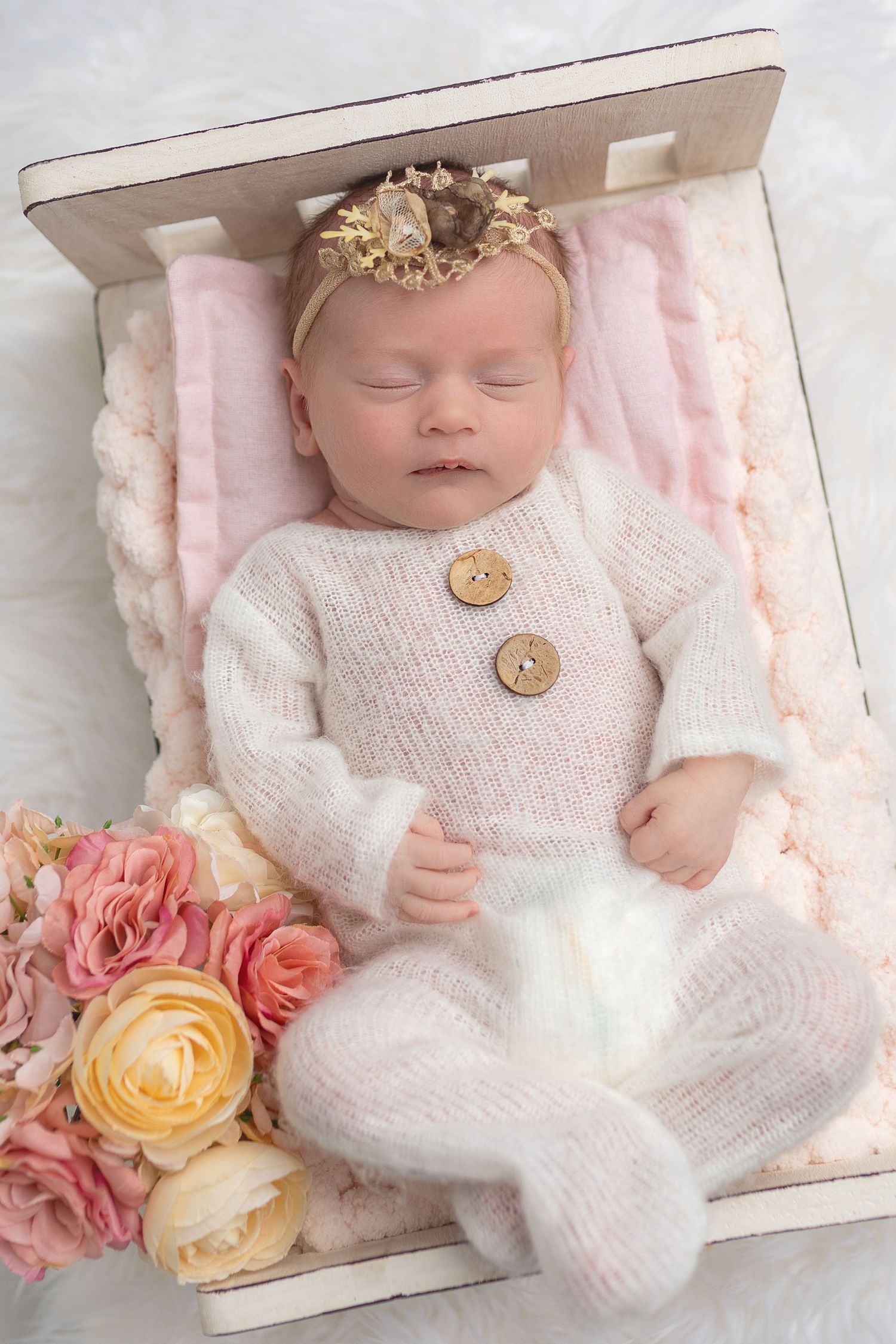 Newborn baby girl sleeping peacefully in knit romper with flower headband on a white miniature bed prop, posed by Cedar Park Photographer