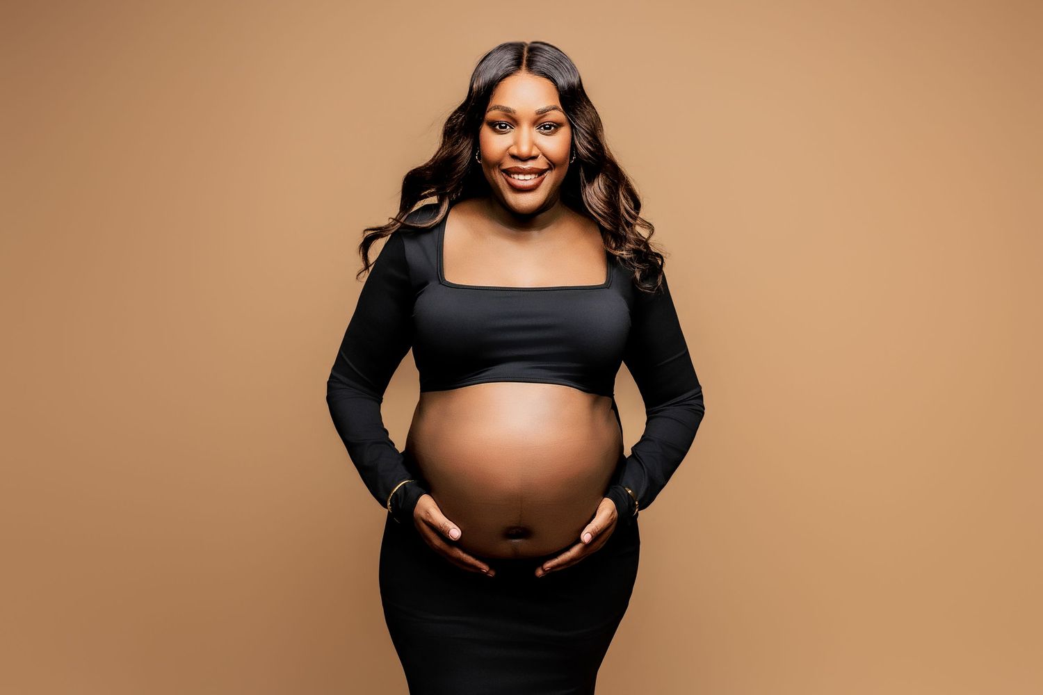 A pregnant woman in a black outfit smiles while cradling her baby bump against a warm brown backdrop.