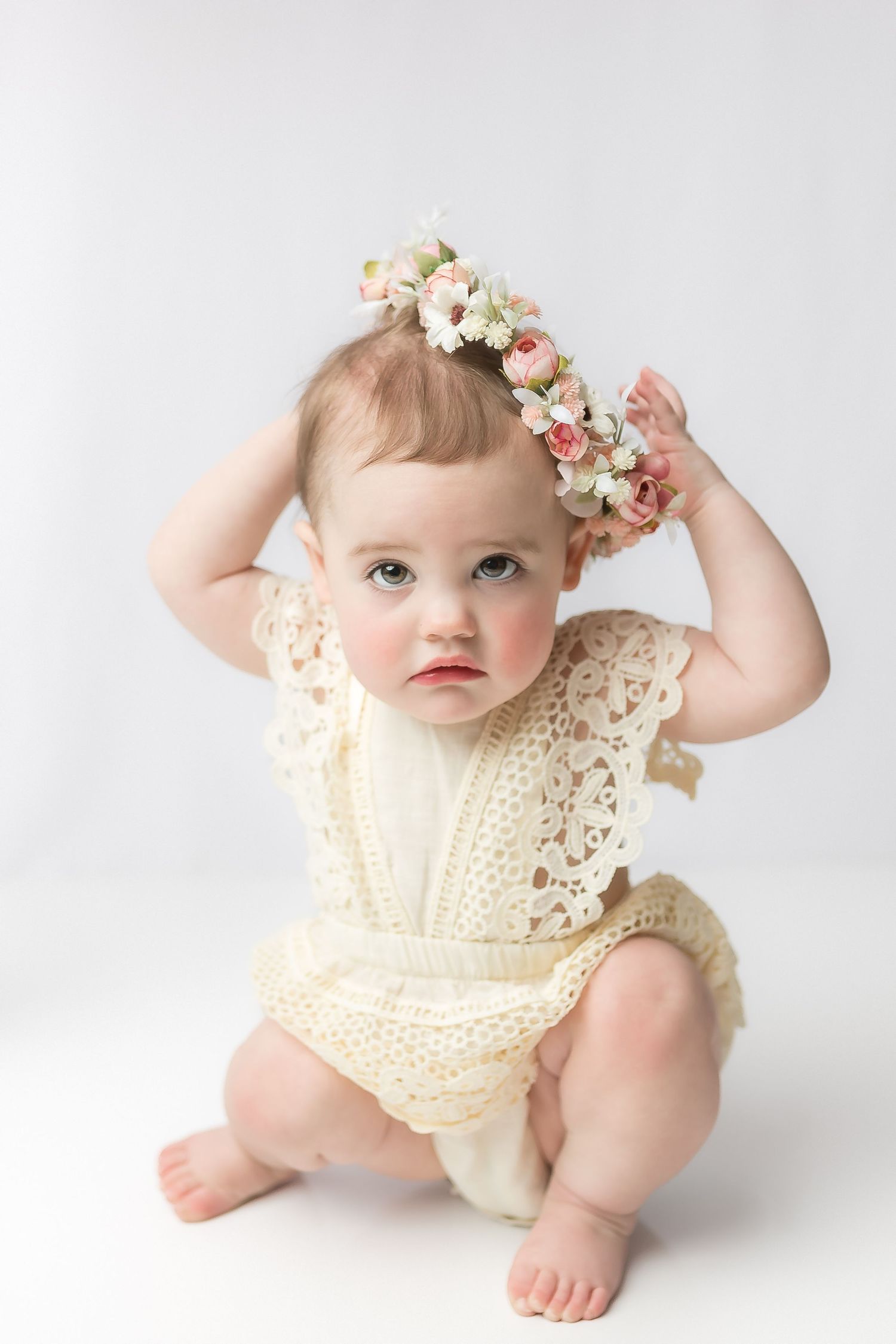 One year old girl stares with big eyes into the lens while holding her flower crown on her head, during her milestone photos in cedar park