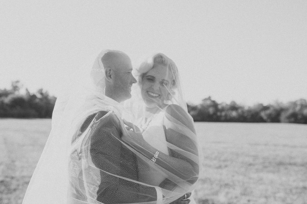 Romantic black and white photo of newlyweds embracing in a sunny meadow with veil flowing in the breeze.