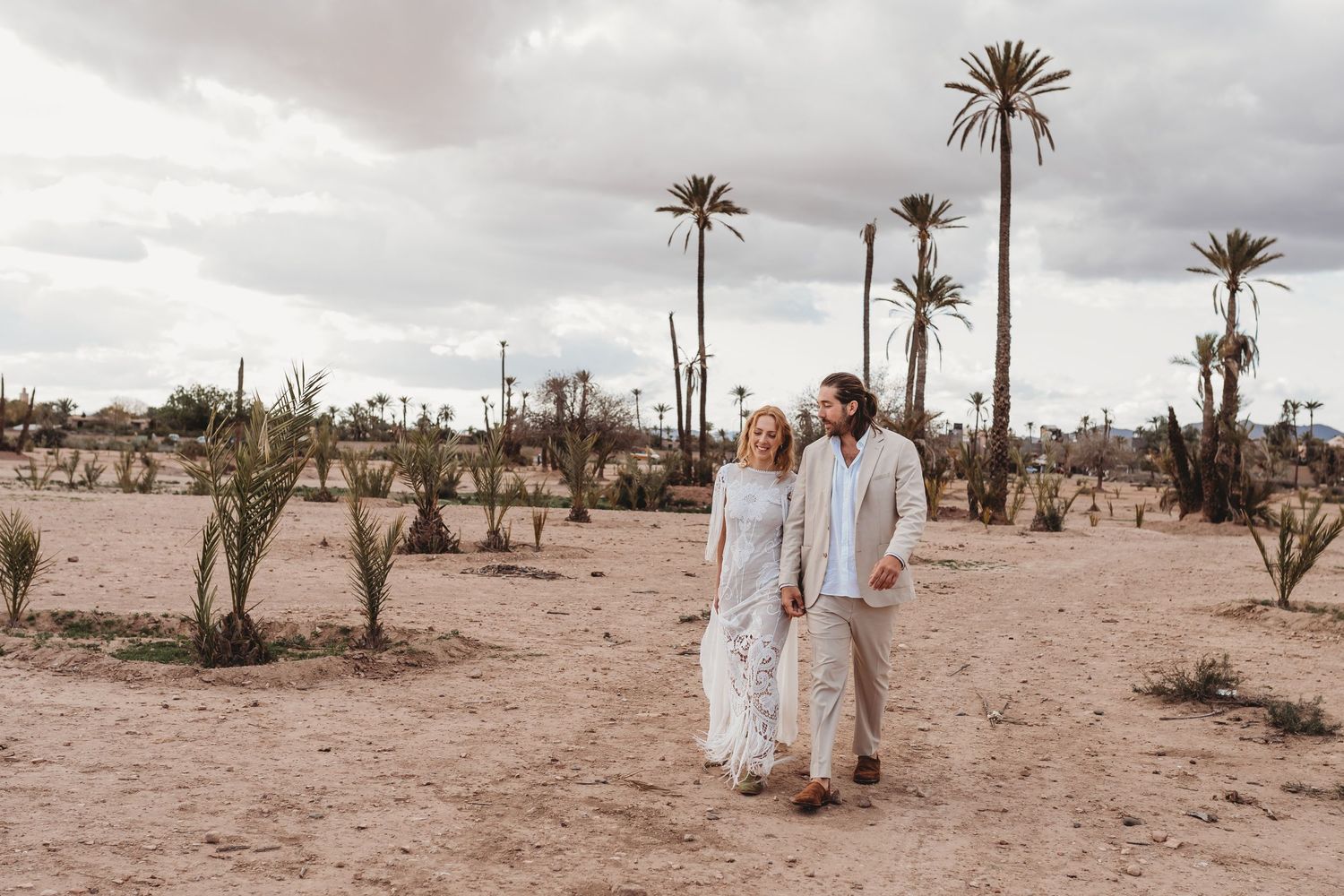 A couple stands in a desert landscape with tall palm trees and cloudy skies in Marrakech, Morocco.