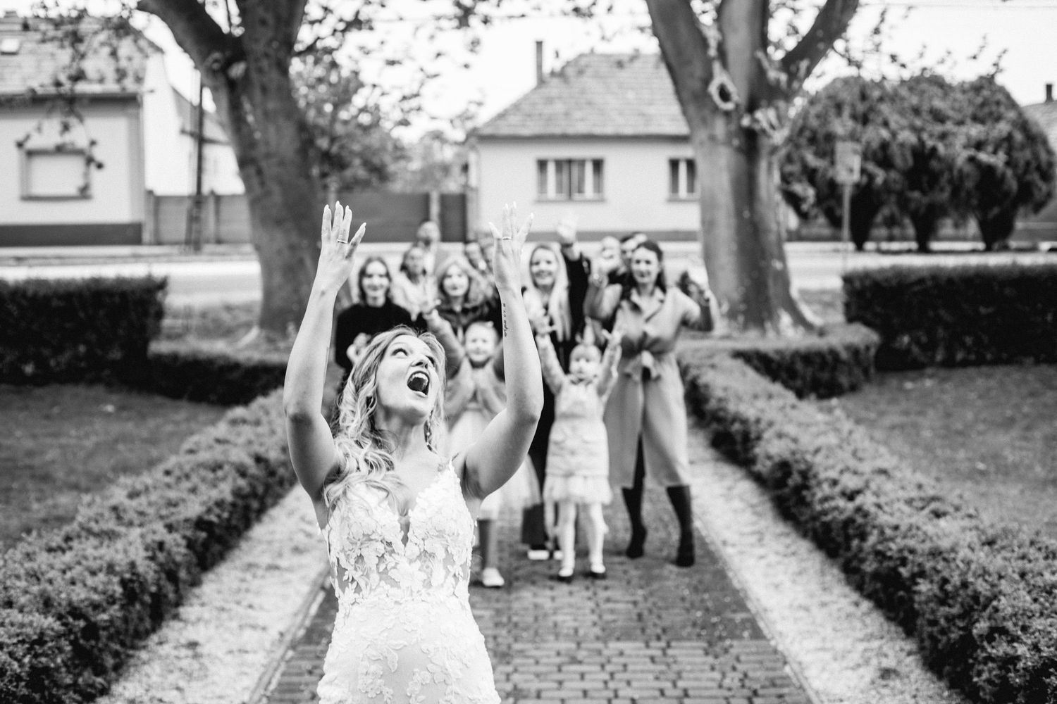 A celebratory moment captured in black and white at an elegant garden venue with people gathered on a tree-lined path.