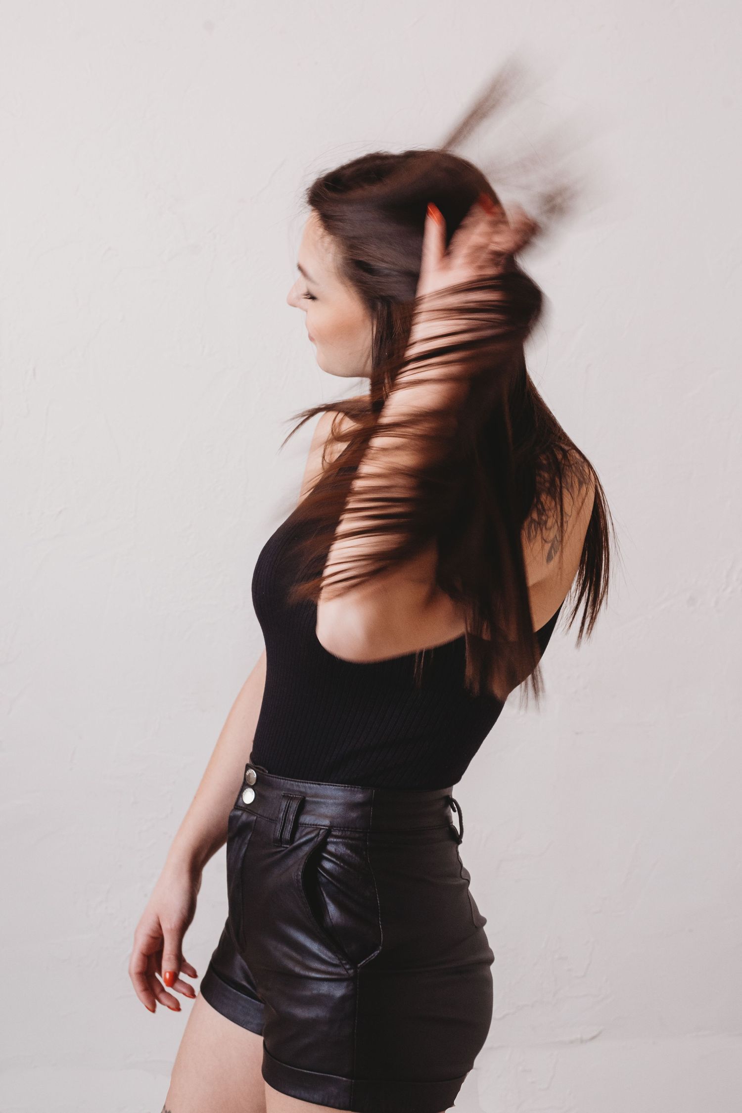 Figure in black top and leather shorts poses dynamically with flowing dark hair against light backdrop.