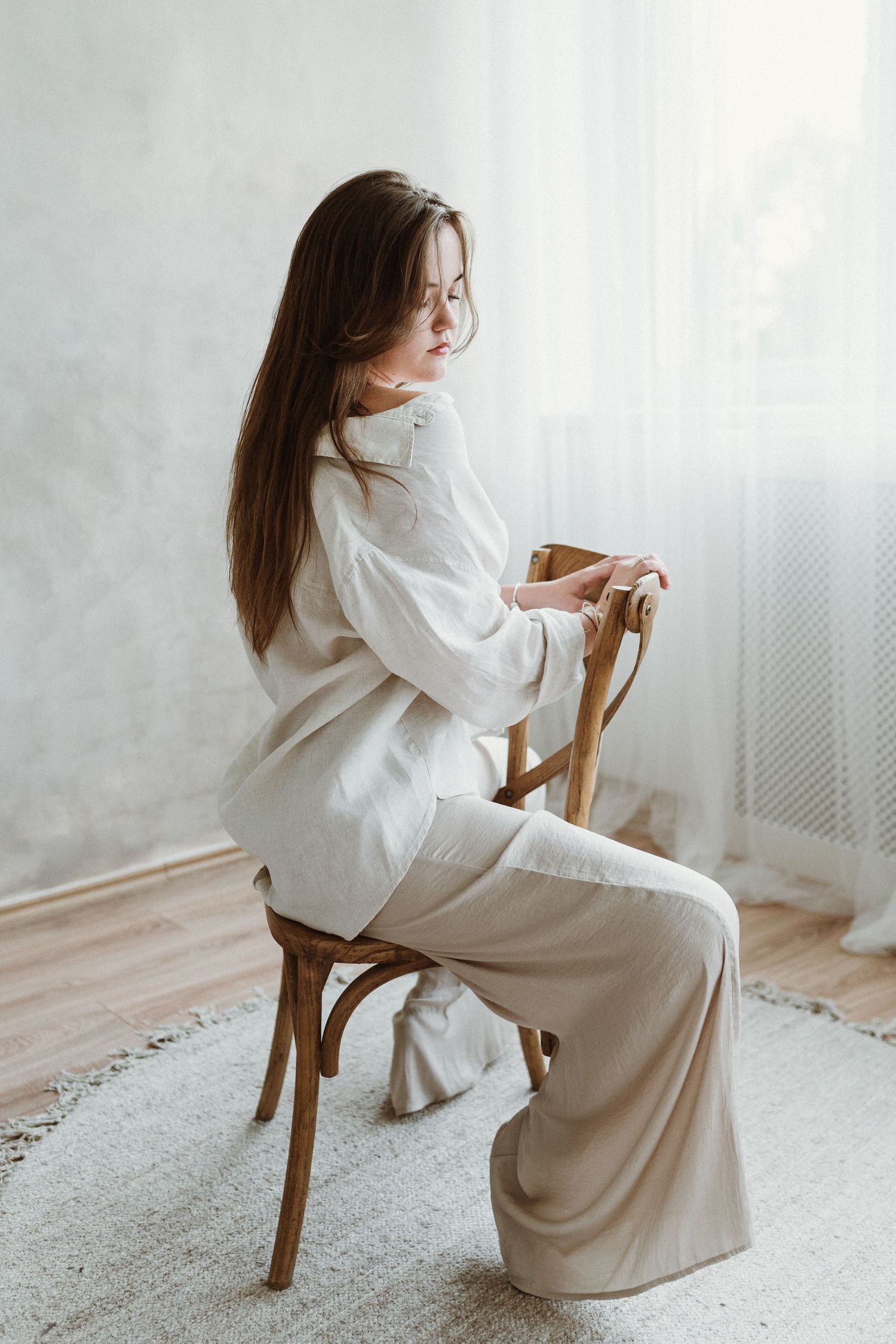 A figure in an elegant white outfit sits sideways on a wooden chair against a light wall with gauzy curtains.
