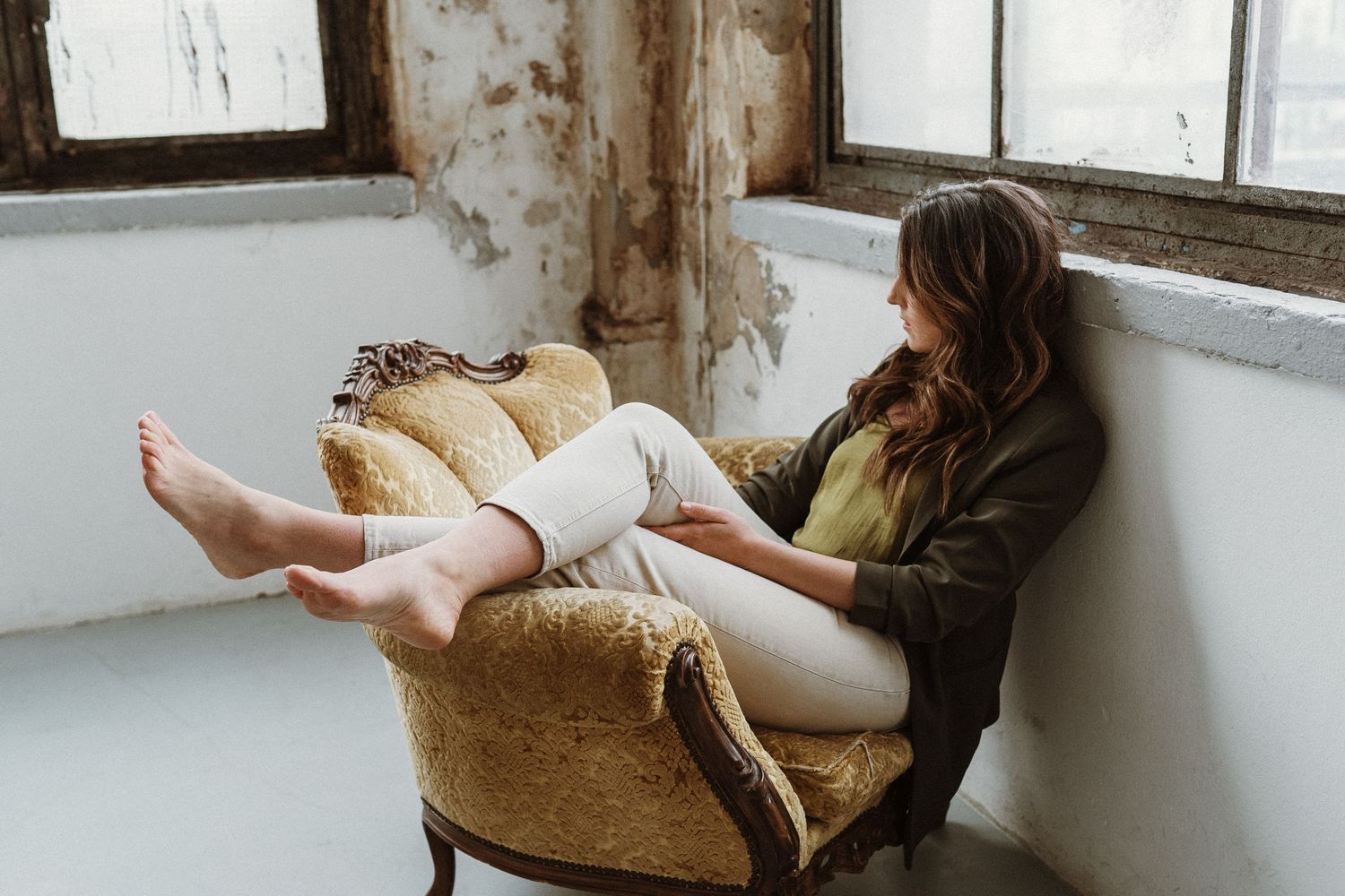 Person relaxes in vintage armchair near window while reading, wearing olive jacket and white pants, feet bare.