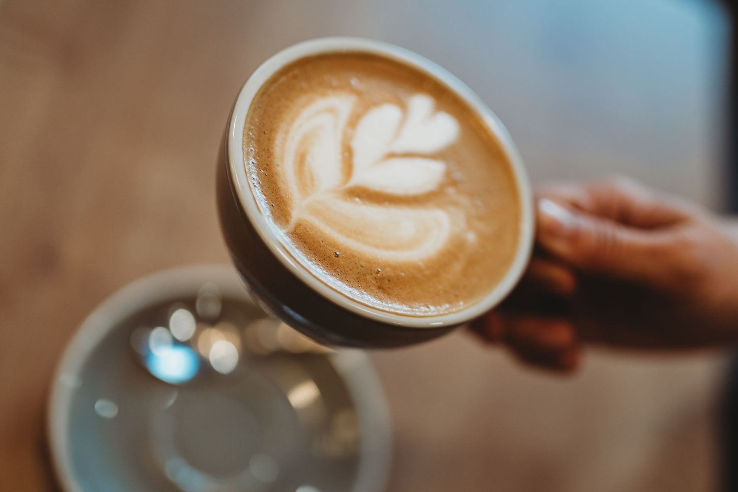 A hand holds a ceramic coffee cup with a delicate latte art flower design against a blurred background.