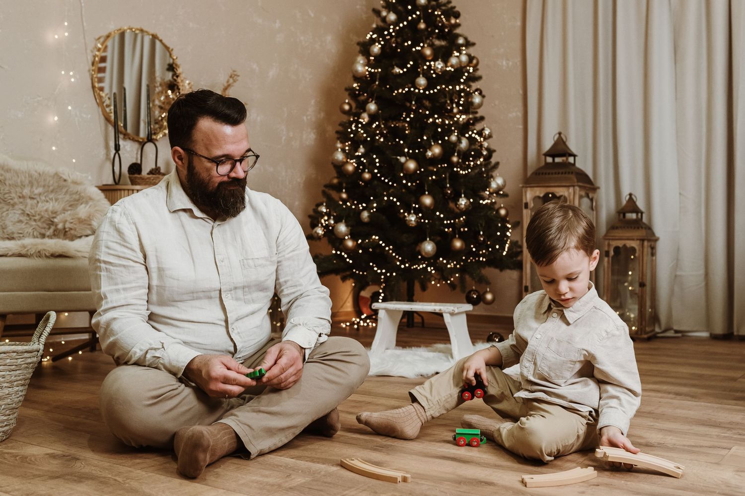 Father and child sit on floor wearing matching beige outfits while playing together near decorated Christmas tree.