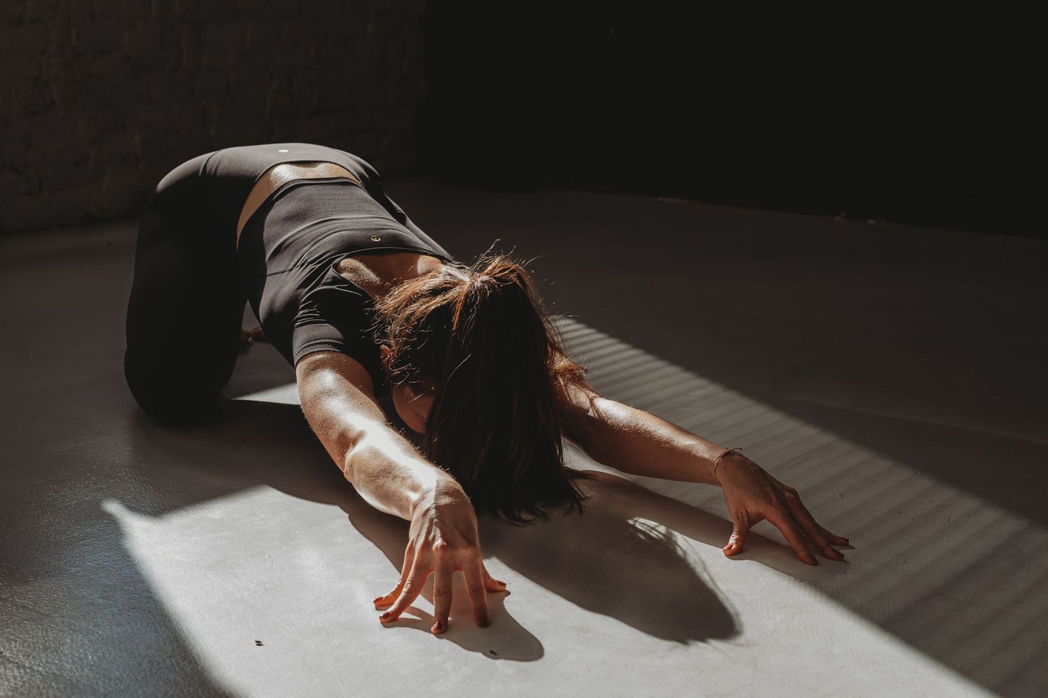 Person stretches on wooden floor in sunlight, creating dramatic shadows in moody artistic composition.
