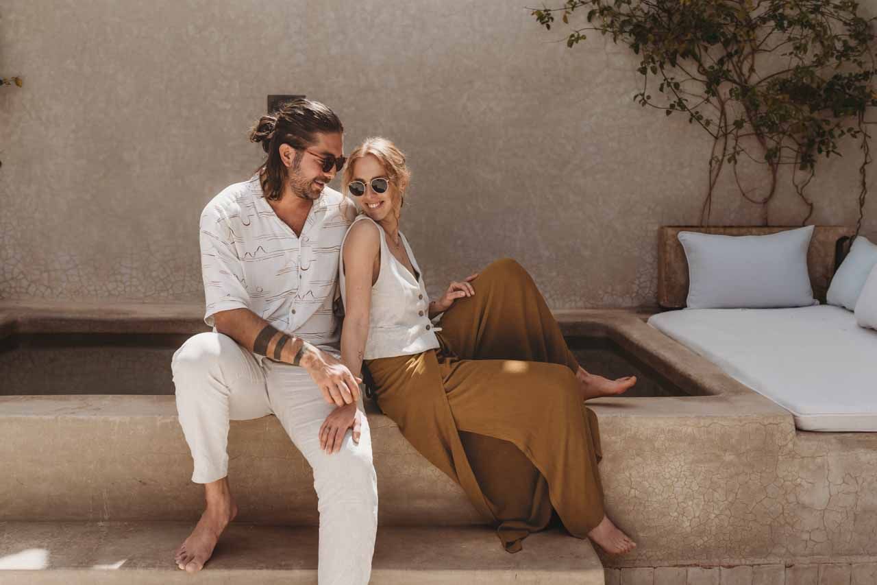 A couple in casual white and brown outfits lounge together on an outdoor patio bench with cushions and plants nearby.