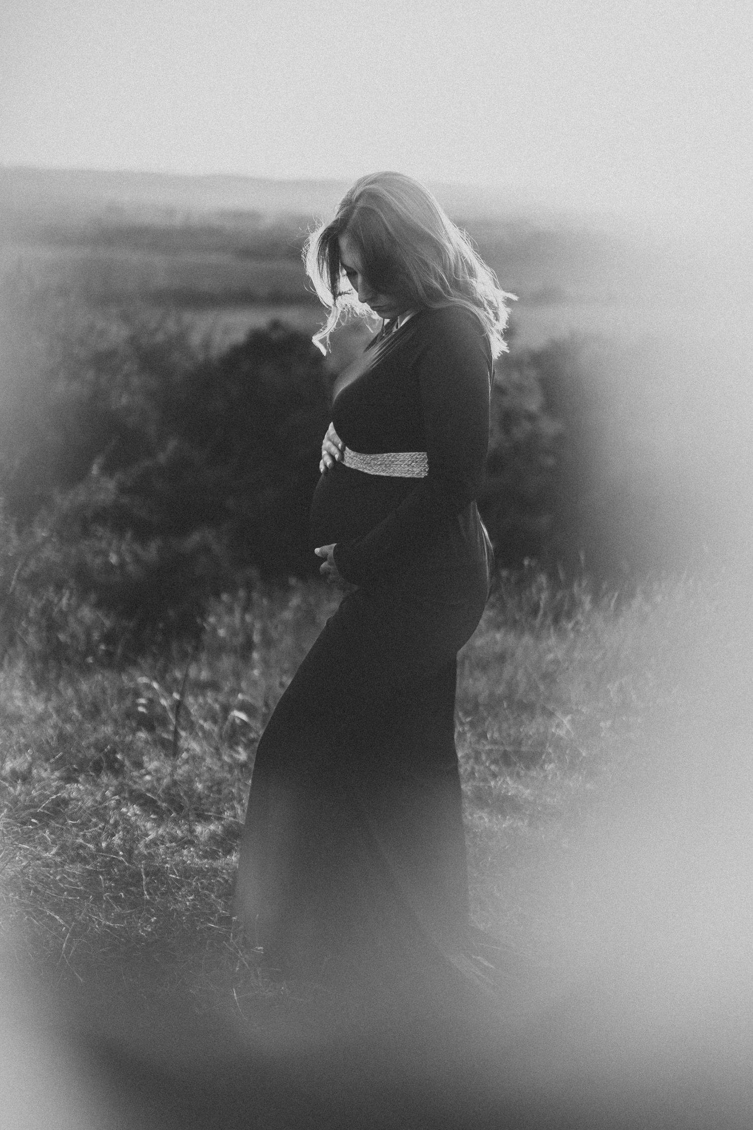 A moody black and white portrait of someone in a dark dress standing in nature with soft lighting and blurred edges.