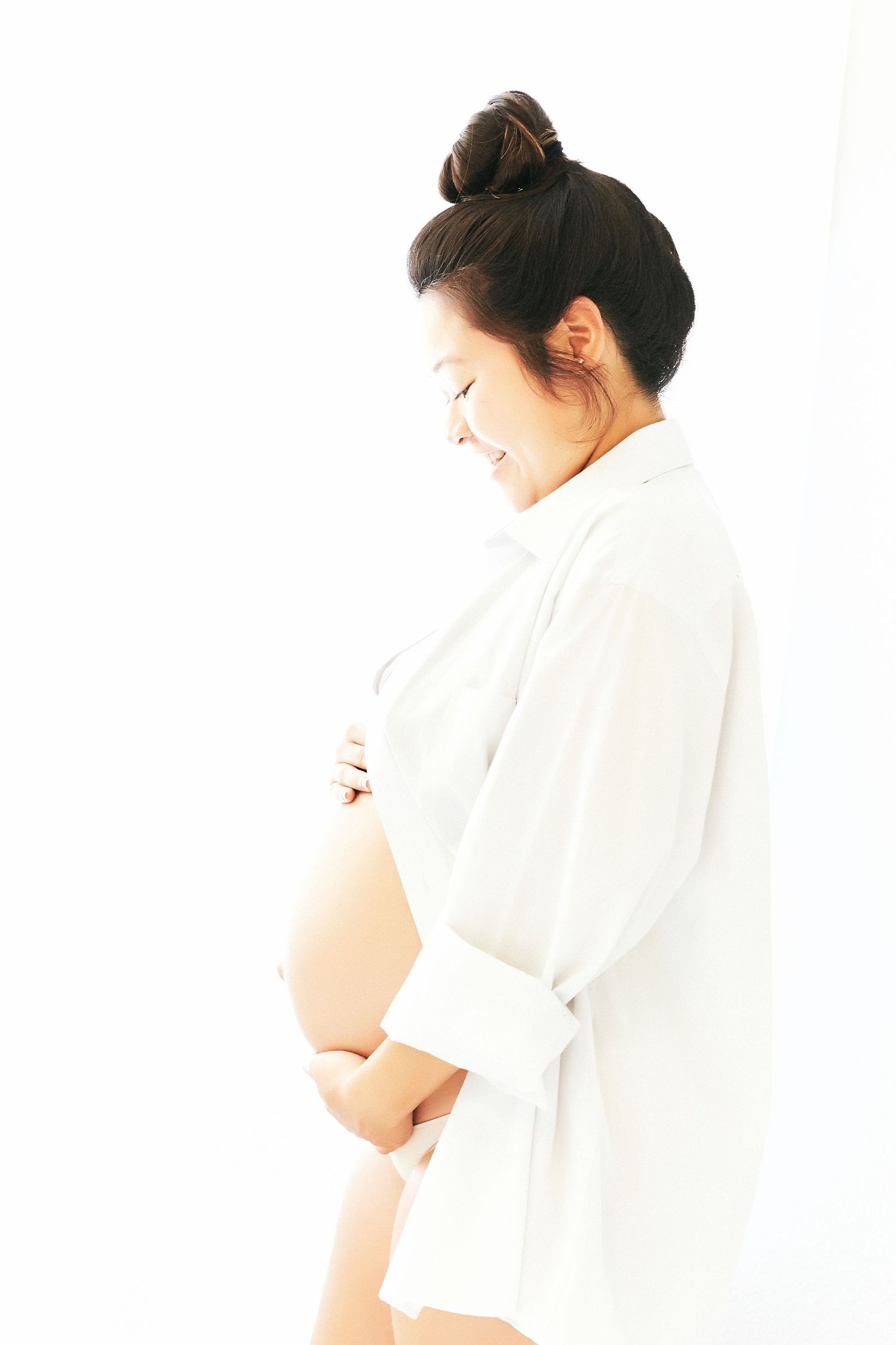 Side profile of expectant mother in white robe holding belly against bright background.