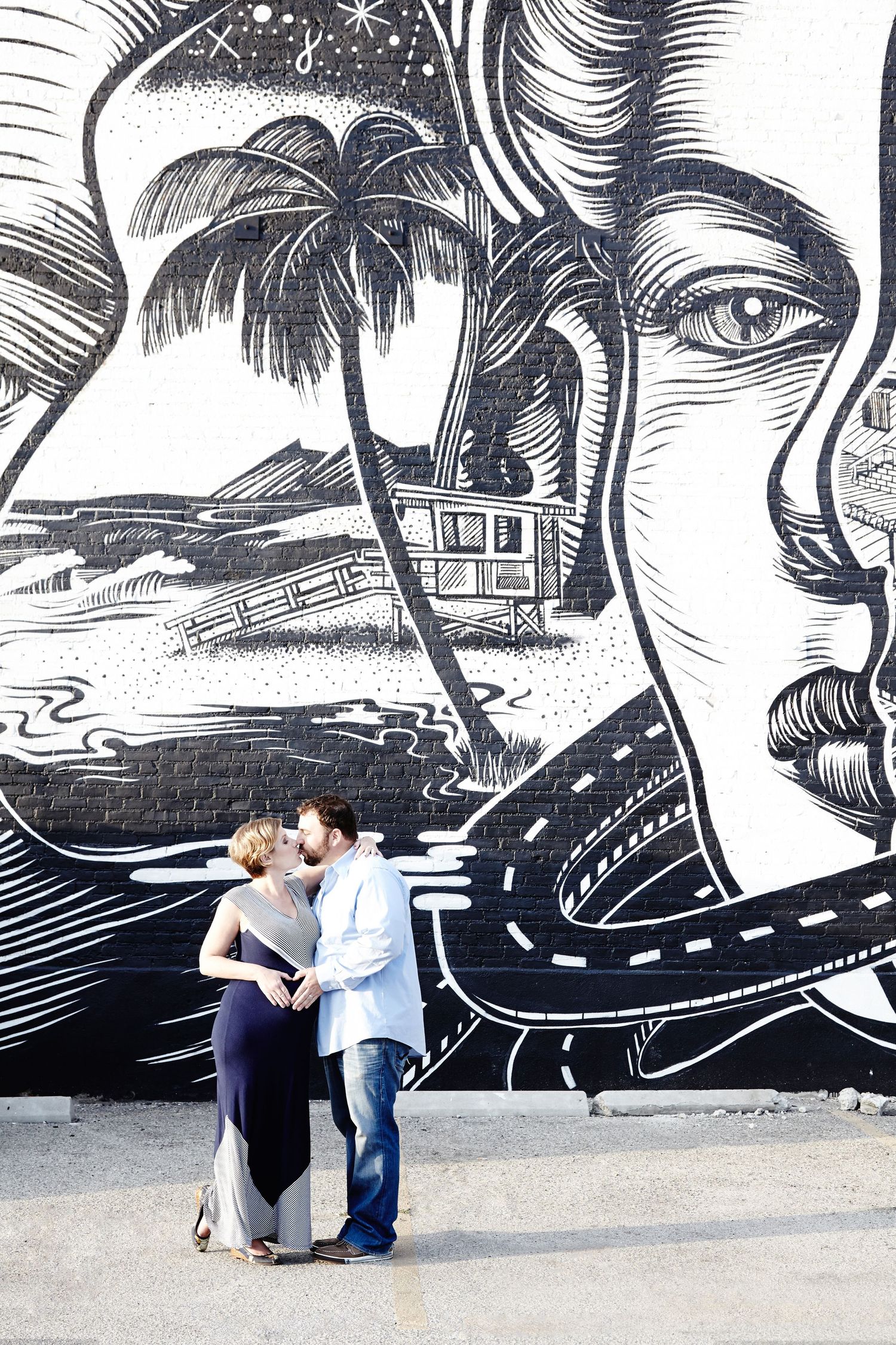 Couple sharing intimate moment against large wall mural featuring palm trees and faces.