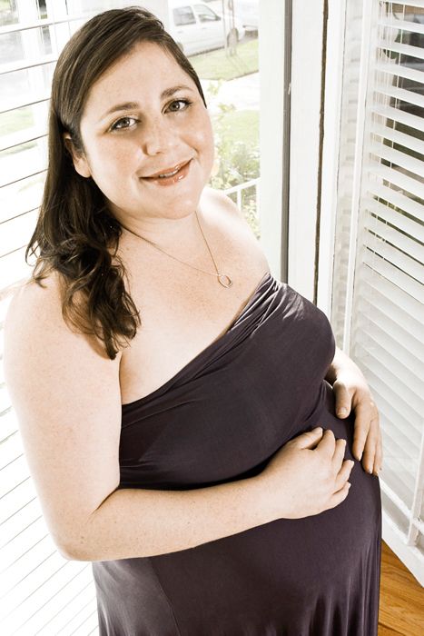 Pregnant woman in dark dress standing by window blinds.