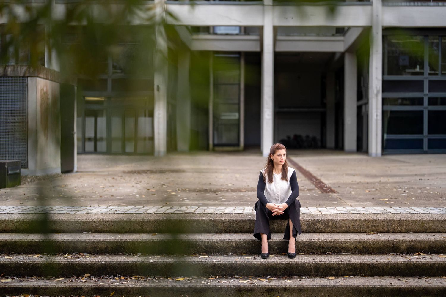 Someone sits alone on concrete steps outside a modern building entrance with glass doors and white columns.