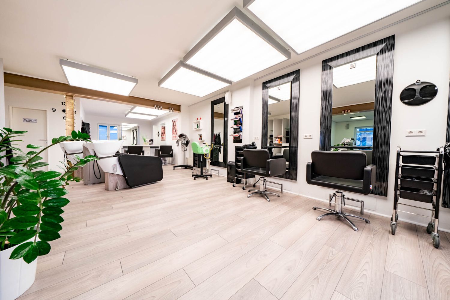 Modern hair salon interior with light wood floors, skylights, and black styling stations in a bright open space.
