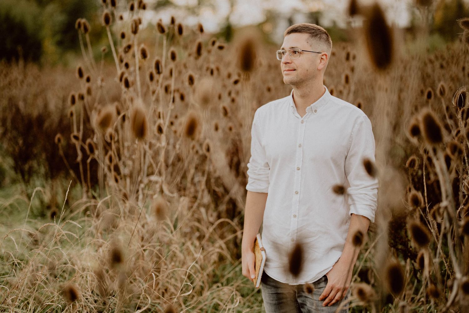 A figure in a white shirt stands among tall brown grass in a natural outdoor setting during golden hour lighting.